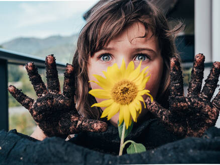 Unbekümmertes Kind mit schmutzigen Händen, das nach vorne gegen eine blühende Sonnenblume auf einem Balkon im Grünen schaut - ADSF31271