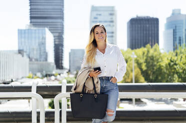 Smiling businesswoman with bag holding mobile phone while leaning on railing - PGF00839