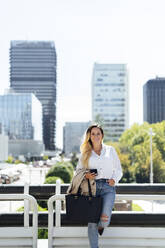 Lächelnde Geschäftsfrau mit Tasche und Mobiltelefon, die sich an ein Geländer lehnt - PGF00841