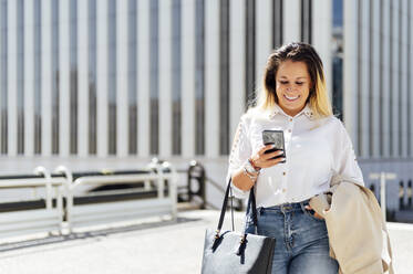 Lächelnde Freiberuflerin mit Tasche Textnachrichten auf Smartphone in der Stadt - PGF00844