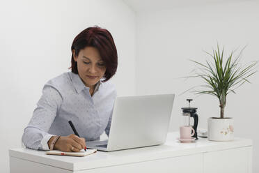 Geschäftsfrau schreibt in Notizblock bei der Arbeit im Büro zu Hause - MOMF00928