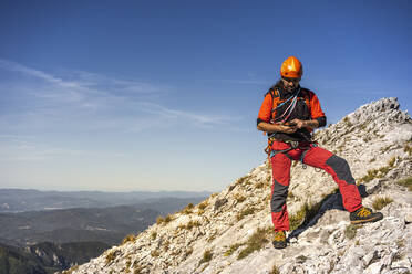 Älterer männlicher Bergsteiger, der sein Smartphone benutzt, während er auf einem Berg vor dem Himmel steht - MCVF00919