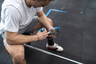 Sportler, der ein Smartphone benutzt und einen Energy-Drink zu sich nimmt, während er auf einer Hantel im Fitnessstudio sitzt - SNF01525