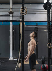 Shirtless male athlete looking at gymnastic rings in gym - SNF01553