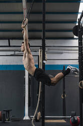 Young male athlete swinging while holding gymnastic rings in gym - SNF01554