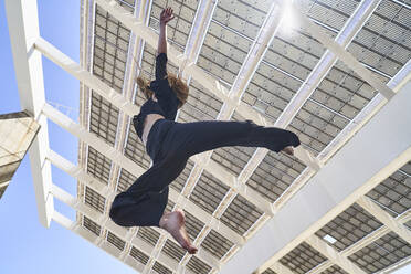 Carefree woman with hand raised jumping in front of building on sunny day - RDGF00357