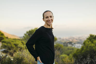 Smiling woman standing with hand in pocket on mountain - DMGF00560