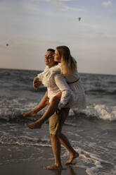 Boyfriend giving girlfriend piggyback ride while walking in water at beach - SSGF00092