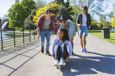 Freunde spielen mit einer Frau, die im Park auf einem Skateboard sitzt - WPEF05464