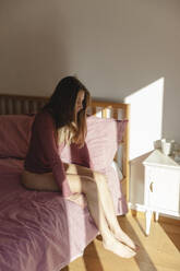 Traurige junge Frau sitzt allein auf dem Bett im Schlafzimmer - MRAF00660