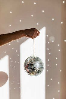 Young woman holding shiny disco ball in front of wall at home - MRAF00667