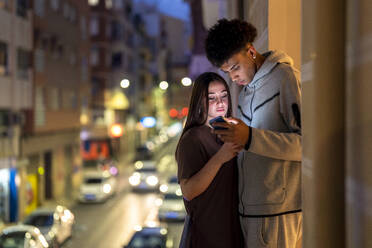 Multiethnisches Paar benutzt sein Smartphone, während es nachts auf dem Balkon steht - DLTSF02339