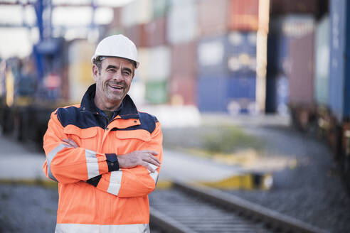 Happy male blue-collar worker with arms crossed in dock - UUF24950