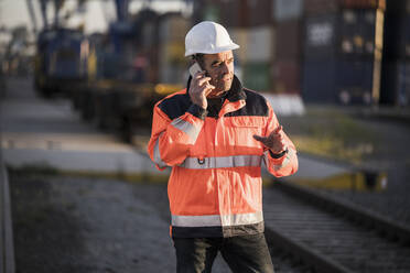Älterer männlicher Hafenarbeiter gestikuliert, während er mit seinem Smartphone an einer Eisenbahnstrecke in der Industrie spricht - UUF24963
