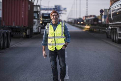 Smiling male truck driver with hand in pocket walking on street at dock - UUF24975