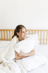 Smiling young woman embracing pillow while sitting on bed in bedroom - MRAF00671