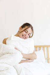 Glückliche junge Frau mit geschlossenen Augen umarmt weißen Kissen zu Hause - MRAF00672