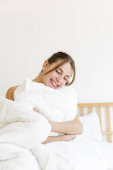 Happy young woman with eyes closed embracing white pillow at home - MRAF00672