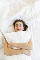 Smiling young woman embracing pillow while sleeping on bed - MRAF00673