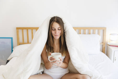 Young woman with brown hair holding cup of coffee sitting on bed - MRAF00688