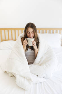 Junge Frau mit geschlossenen Augen trinkt Kaffee, während sie zu Hause in eine Decke eingewickelt ist - MRAF00690