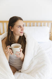 Junge Frau hält eine Kaffeetasse, während sie zu Hause in eine weiße Decke eingewickelt ist - MRAF00692