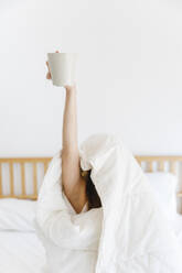 Frau mit erhobener Hand, die eine Kaffeetasse hält, eingewickelt in eine weiße Decke auf dem Bett - MRAF00693