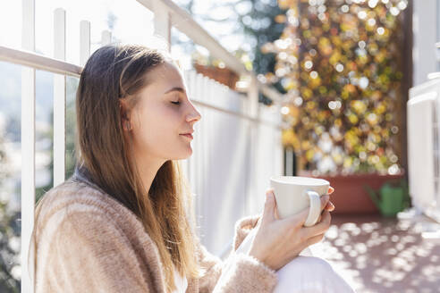 Young woman holding coffee cup while sitting with eyes closed in balcony - MRAF00694