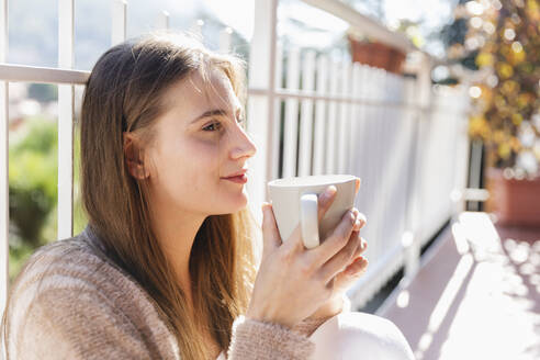Junge Frau mit Kaffeetasse, die auf einem Balkon sitzt und nachdenkt - MRAF00695
