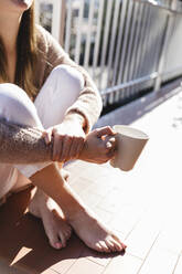 Junge Frau sitzt mit Kaffeetasse auf dem Balkon - MRAF00696