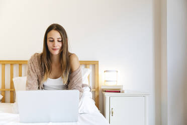 Junge Frau benutzt einen Laptop, während sie zu Hause auf dem Bett sitzt - MRAF00699