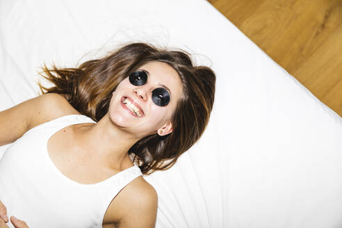Glückliche junge Frau, die sich die Augen mit Lakritzbonbons bedeckt, während sie zu Hause auf dem Bett liegt - MRAF00704