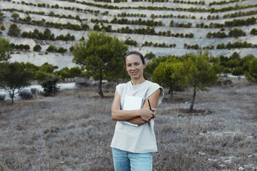 Smiling scientist standing with book and pen in agricultural field - DMGF00595