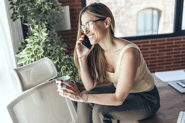 Smiling female freelancer with coffee mug talking on smart phone - JSRF01670