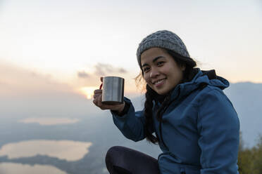Smiling woman showing coffee mug on sunset during vacation - MRAF00740