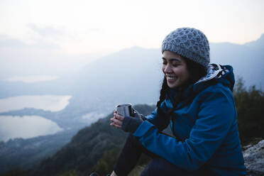 Lächelnde Frau mit geschlossenen Augen, die einen Kaffeebecher auf einem Berg hält - MRAF00744