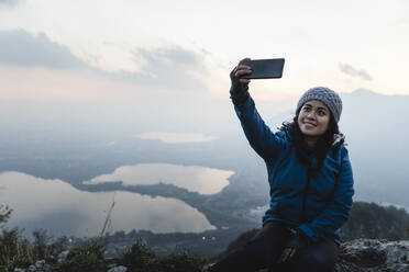 Lächelnde junge Frau nimmt Selfie durch Handy auf Sonnenuntergang - MRAF00749