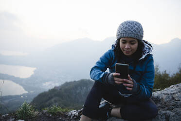 Wanderer mit Strickmütze, der auf einem Berg ein Mobiltelefon benutzt - MRAF00750