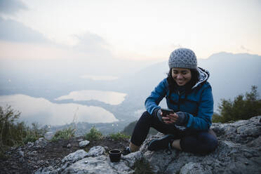 Glückliche junge Frau, die auf einem Berg eine Textnachricht über ihr Smartphone verschickt - MRAF00752