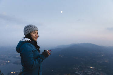 Lächelnde junge Frau mit Kaffeebecher auf einem Berg im Urlaub - MRAF00753