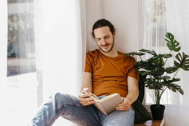 Man reading book while sitting at window - TCEF02088