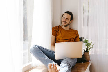 Smiling man with laptop sitting at window - TCEF02089