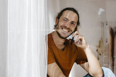 Lächelnder Mann, der mit seinem Handy telefoniert und aus dem Fenster schaut - TCEF02093