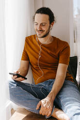 Mann mit Mobiltelefon hört zu Hause Musik über In-Ear-Kopfhörer - TCEF02094