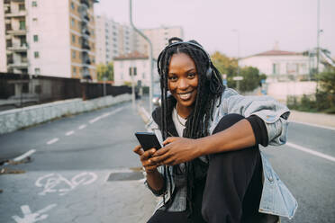 Lächelnde Frau mit Mobiltelefon, die auf dem Fußweg Musik hört - MEUF04614