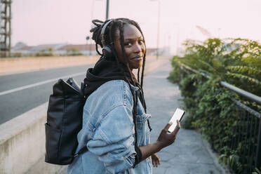 Smiling woman with backpack holding mobile phone on footpath - MEUF04617