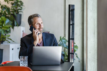 Businessman with laptop talking on smart phone in office - DIGF16595