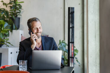 Smiling businessman using laptop while talking on phone in office - DIGF16596