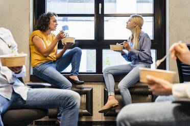 Businesswomen having food at window sill with colleagues in office - DIGF16691