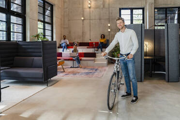 Lächelnder Geschäftsmann mit Fahrrad im Büro stehend - DIGF16781